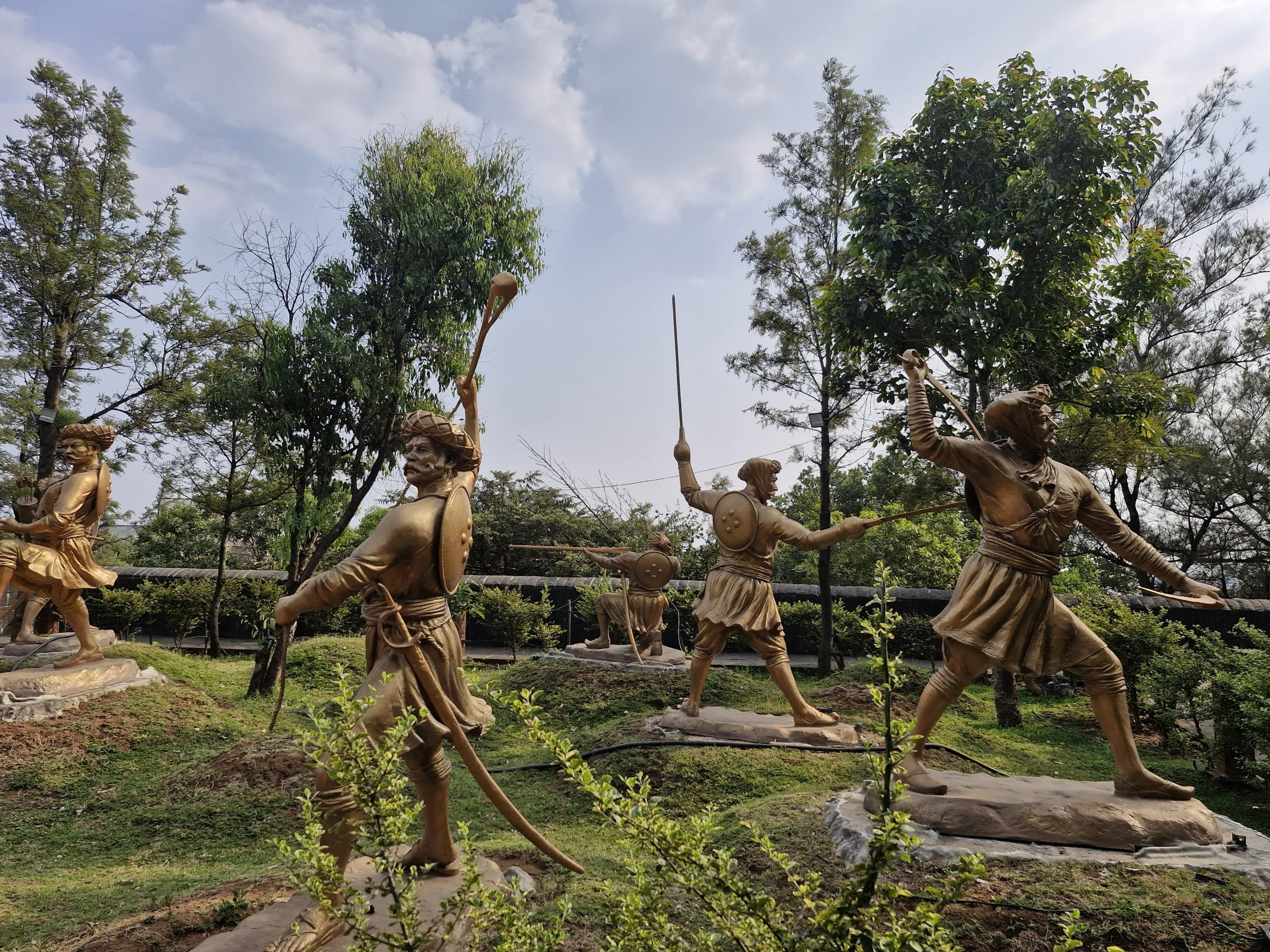 Golden Maratha warrior statues mid-charge in the memorial garden at Sinhagad Fort, with the Sahyadri treeline behind them.